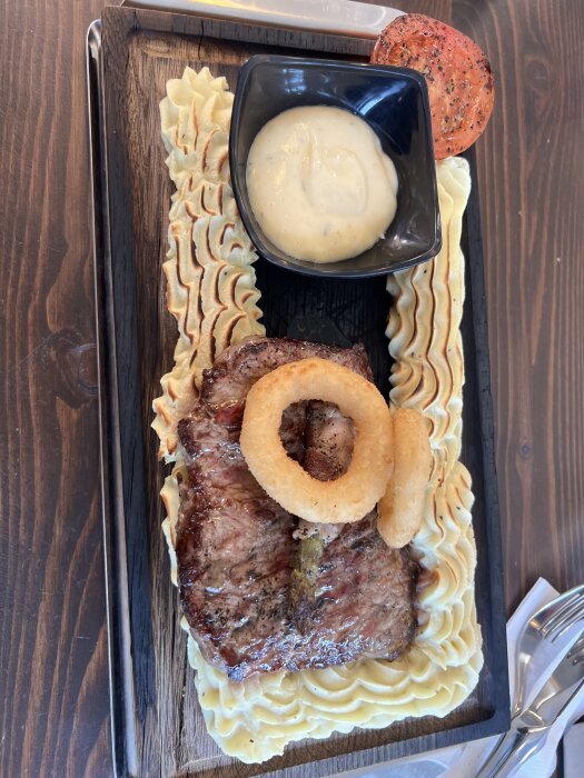 Stek med lökringar, mos runt tallriken, sås i skål, och en grillad tomat.