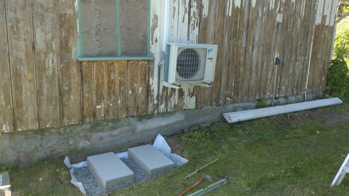 Gammal träfasad med flagnande färg på sommarstuga. Luftvärmepump installerad. Förberedelse för utomhusprojekt pågår.