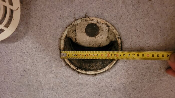 Golvbrunn i dusch med synliga sprickor, omgiven av grå kakel, där ett måttband visar diametern och en hand håller måttbandet.