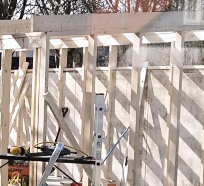 Bygge av träregelvägg med verktyg och stege framför, reglar på betongvägg med cellplastremsa emellan.