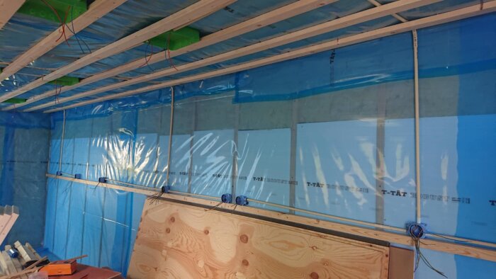 Isolerat rum med blå diffusionsspärr, träreglar i taket, och kablar för elinstallation över plywoodskiva vid betongvägg.