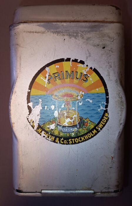 Sliten etikett på en gammal förvaringsask med texten "Primus" och en illustration av en bensinkök, omgiven av färgglada solstrålar och landskap.
