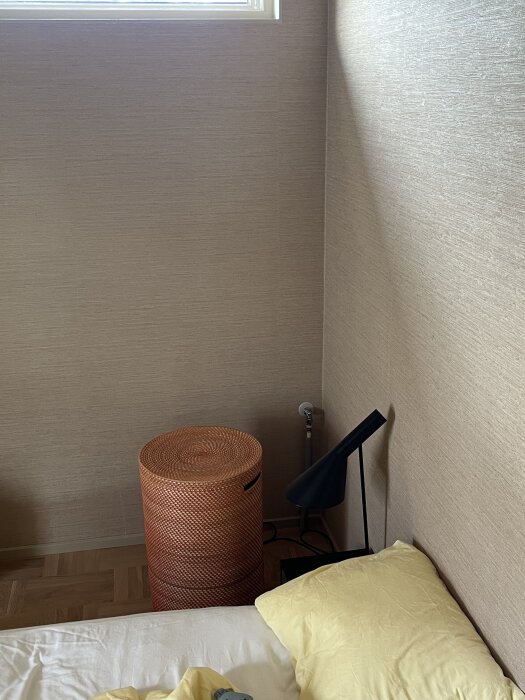 Hörn av sovrum med neutral tapet, en orange flätad korg, svart lampa och gul kudde. Synliga rör vid golv.