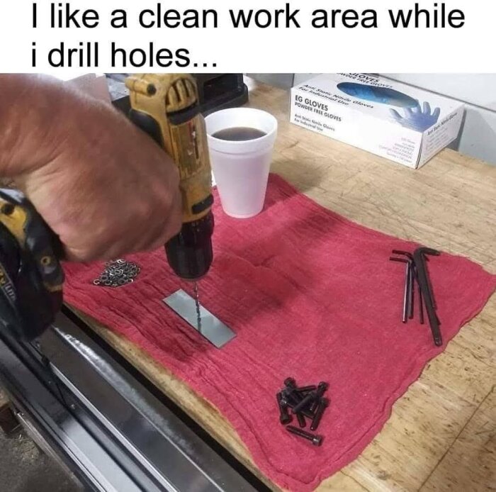 En person borrar i metall med en borrmaskin på en arbetsbänk, med skruvar, insexnycklar och en kaffemugg på en röd trasa.