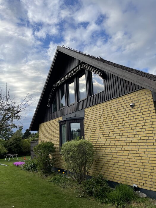 Gult tegelhus från 1973 med svart träpanel och flera fönster i takt med en trädgård i förgrunden.