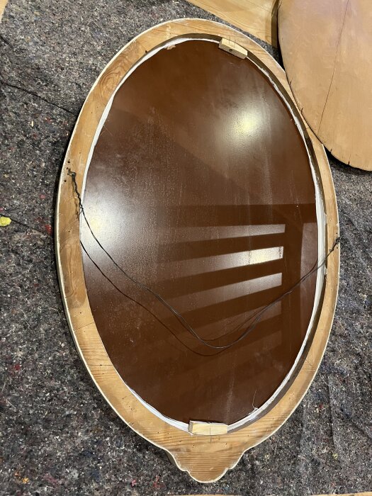 Oval spegel med träram och fog, två träbitar på baksidan för stabilitet, placerad på filtmatta efter renoveringsprojekt.