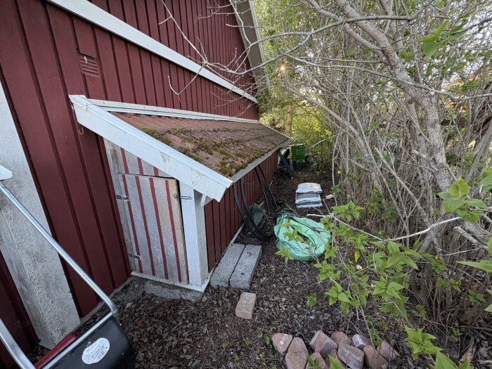 Förvaringsutrymme bakom rött garage med grönska, där ved och andra föremål som säckar och tegelstenar är ordnade för förbättrad förvaring.