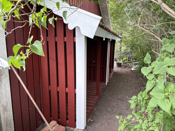 Rött förvaringsutrymme bakom garaget med plats för ved, omgivet av träd och buskar.