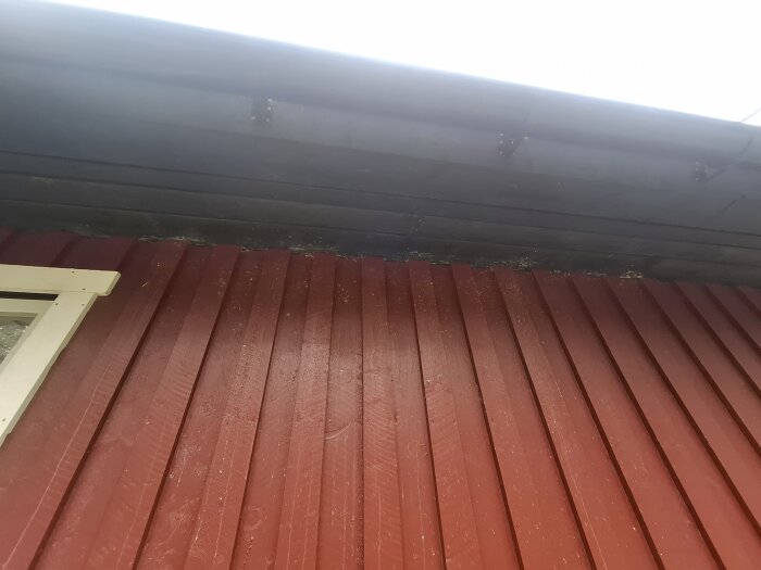 Röd träpanelvägg med ett svart plåttak ovanför, där takfoten möter väggen; ser märkligt ut med potentiella skador.