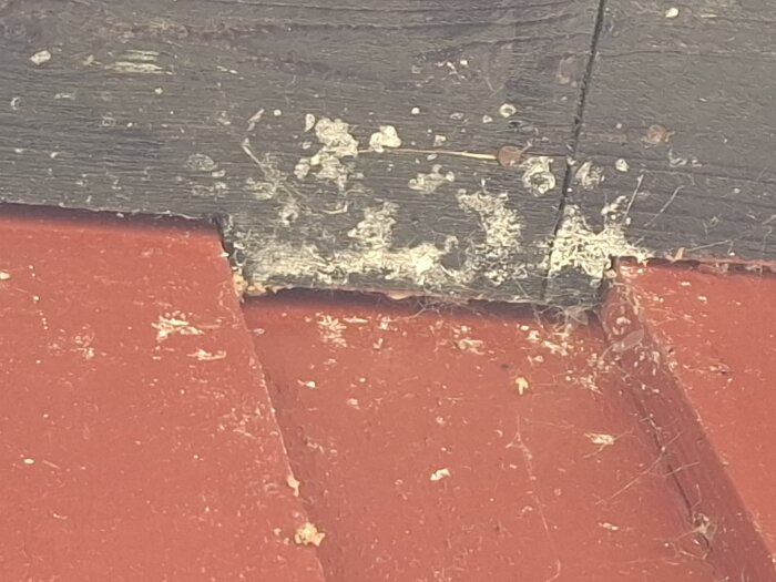 Närbild på takfot där rött plåttak möter svart fiberskiva, med vita fläckar och spår av smuts och damm mellan materialen.