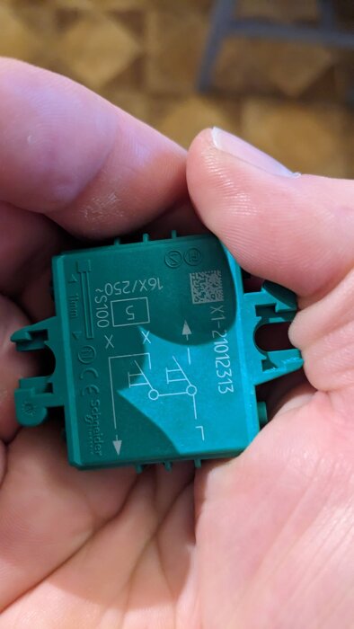 Grön komponent med elektriska symboler och text, inklusive "Schneider". Hålls i handen, detaljer synliga.