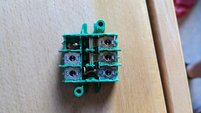 Grön plastmontering av strömställare på träbord, med metalldelar synliga, demonterad och vänd uppåt.