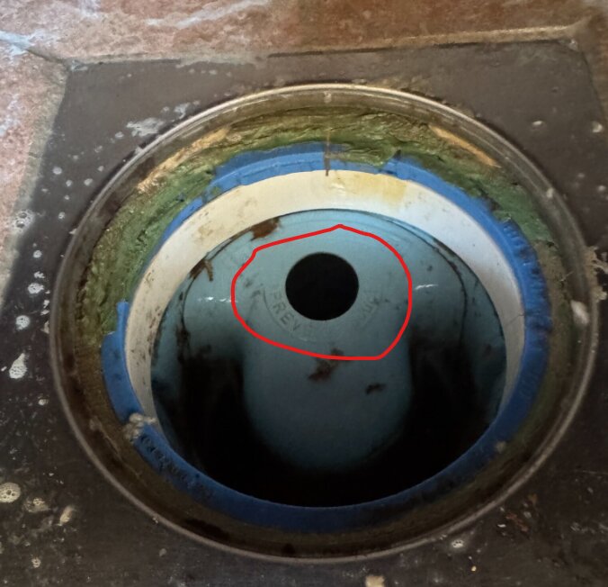 En avloppsbrunn med borttagen propp, omgiven av smuts och grön beläggning på kanten. Röd markering runt hålet där proppen suttit.