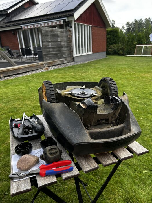 En robotgräsklippare på ett bord med diverse handverktyg för service, inklusive skruvmejslar och tänger, framför ett hus med trädgård.