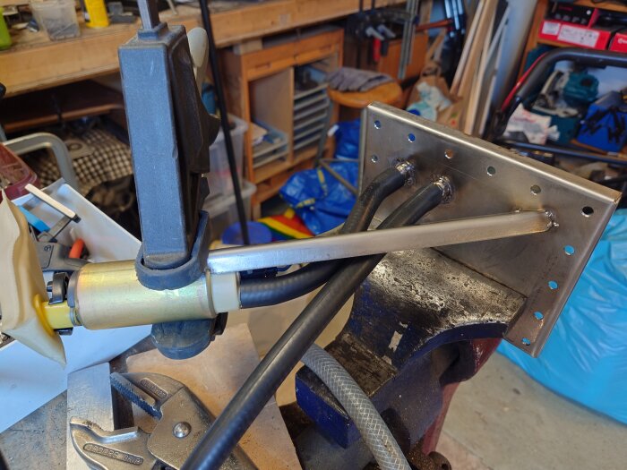 En plåtbit med svetsat fäste, omgiven av rör, fastsatt i ett skruvstäd; hål för attributgenomföringar synliga. Verktyg och material i bakgrunden.