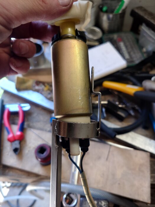 Hand håller i en komponent, kanske en bränslepump, med fäste och kablar, placerad på en arbetsbänk med verktyg i bakgrunden.