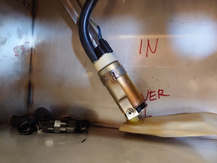 En svetsad fästanordning med monterad sensor och slangar, omgiven av texten "IN" och "OVER", på metallbakgrund.