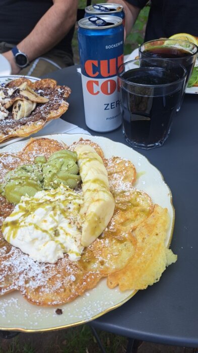 Våffla toppad med banan, grädde och gröna pålägg, serverad med läsk och glas vid sidan på ett bord.