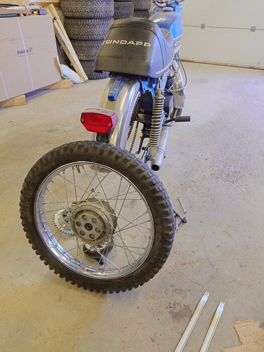 Zündapp-moped utan monterat bakhjul i ett garage, med verktyg och däck staplade i bakgrunden.