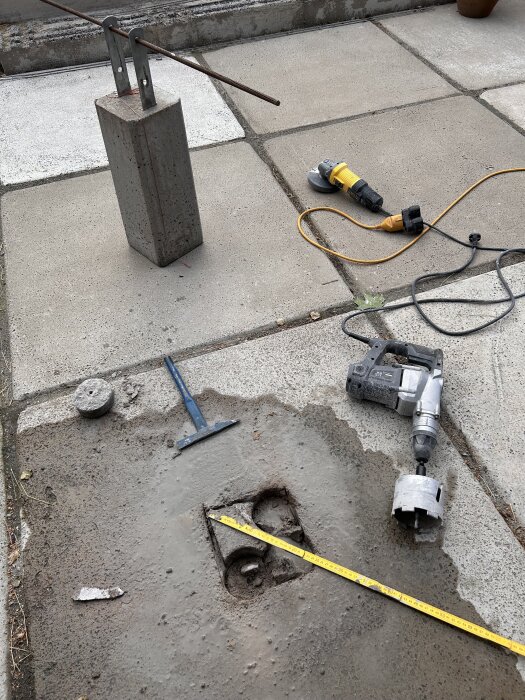 Betongblock med armeringsjärn, borrmaskin och vinkelslip vid ett halvfärdigt fyrkantigt hål på ett betonggolv, mätt med tumstock.