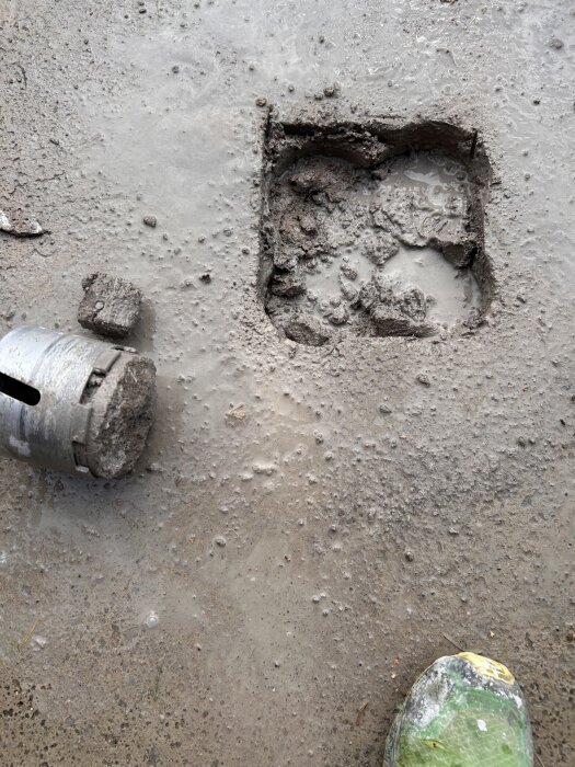 Kärnborr bredvid ett fyrkantigt hål i betonggolv, grävt delvis upp, med fuktig betong runtom och en skyddssko i hörnet av bilden.