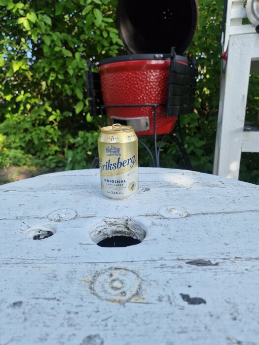 En ölburk står på ett slitet träbord framför en röd kolgrill i en grönskande trädgård.