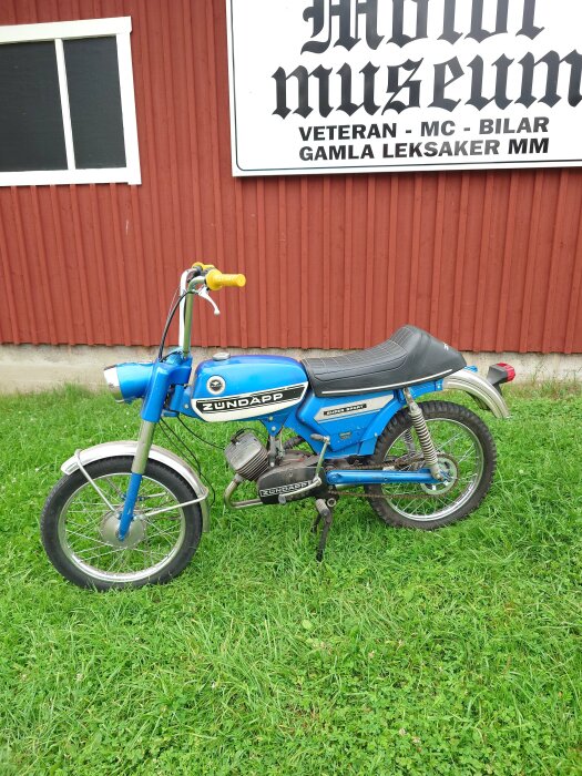 Blå Zündapp-moped parkerad på gräs framför en röd byggnad med skylt för motor- och leksaksmuseum.
