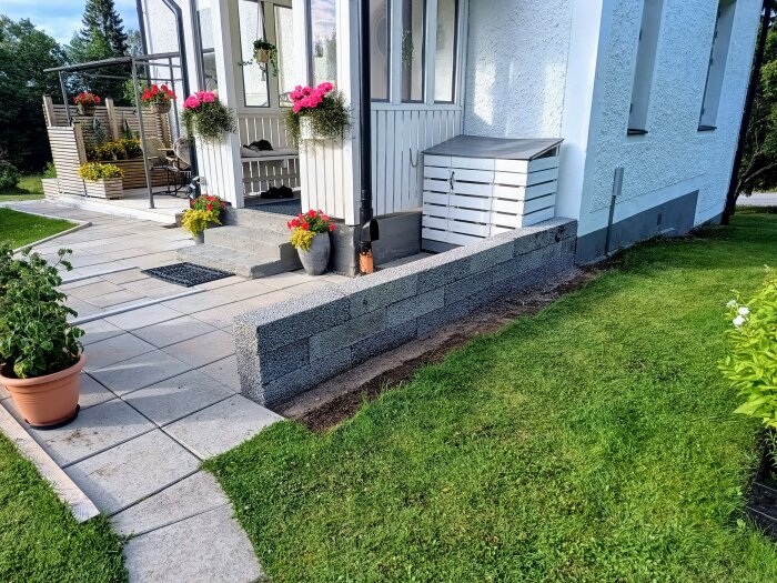 Lecablock staplade till en låg mur bredvid ett hus med vit putsfasad, flera blommor i krukor och en gräsmatta i förgrunden.