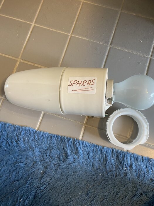 Trasig äldre armatur från 60-talsbadrum med märkt kupglas och lampfot på blå matta och grått kakelgolv.