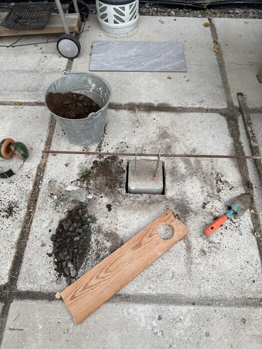 En betongplatta med en hink jord, träbit och trädgårdsverktyg, förberett för ett byggprojekt eller renovering.