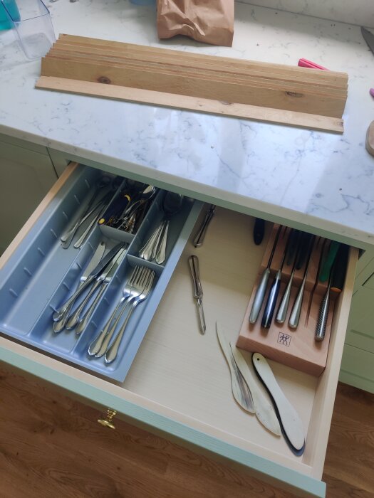 Öppen kökslåda med egengjord bestickinsats av plywood; verktyg och köksredskap organiserade i fack.