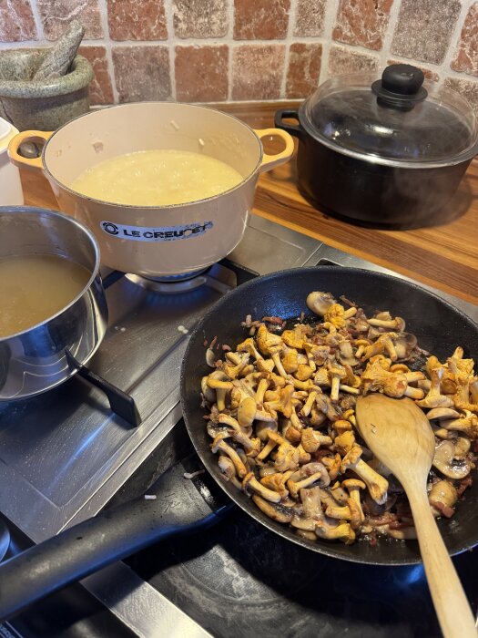 Kantareller och pancetta steks i en stekpanna, bredvid en gryta med risotto och en kastrull med kycklingfond på en spis.