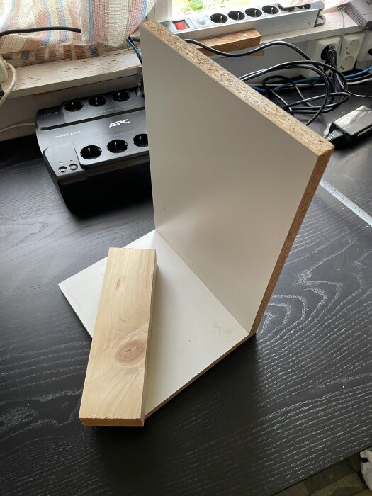 Träbit och två melaminskivor på svart bord framför eluttag och strömsladdar i en verkstadsmiljö.