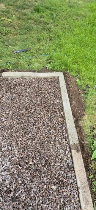 Bilden visar en nygjuten gräskant i betong invid en yta med stenar, med gräs och en hammare i bakgrunden.