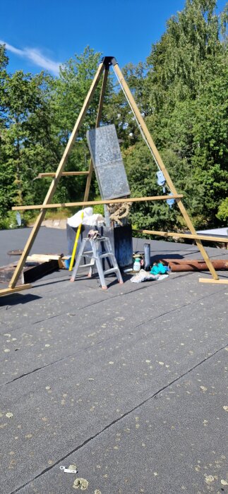 Ställning med plåttrumma och verktyg på tak, omgiven av grönska. Ventilationsrör, stege och arbetstillbehör synliga.