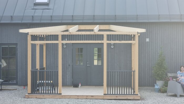 Takstolar monterade på en nybyggd altan framför ett hus, med en person som sitter på sidan intill planterade växter och trädgårdsdekorationer.