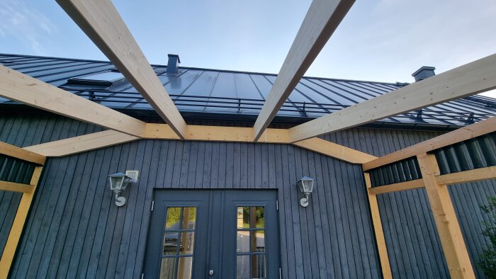 Prefabricerade takstolar installerade på ett hus med mörk träfasad och metalltak, två utelampor vid dörren.