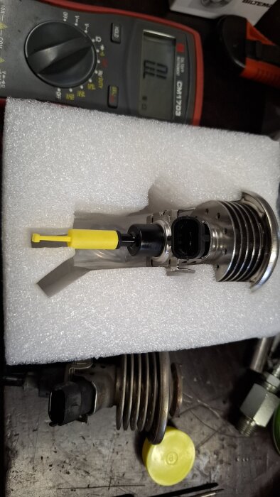 VW Sharan Adblue dosermunstycke ligger på skumgummi, bredvid en multimeter och andra verktyg på en arbetsbänk.