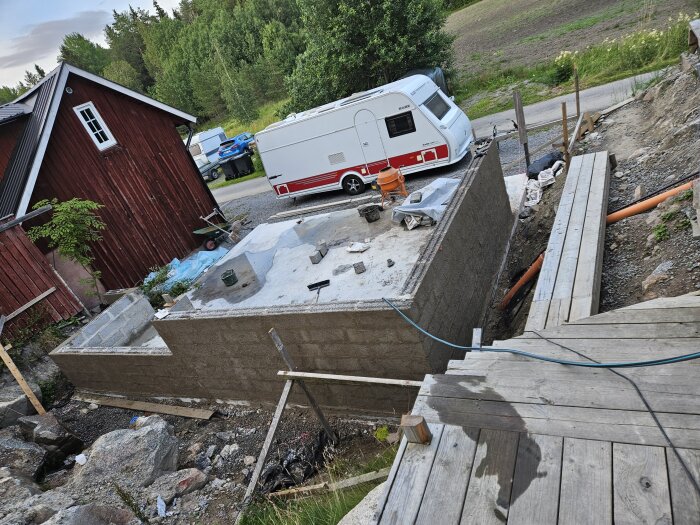 Byggarbetsplats med nyligen murad och slamrad vägg, omgiven av träkonstruktion, röd byggnad och parkerad husvagn i bakgrunden.