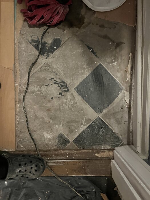 Trägolv med svartvita kakelplattor synliga bland rester av golvspackel. En röd trasa och en svart sko syns också.