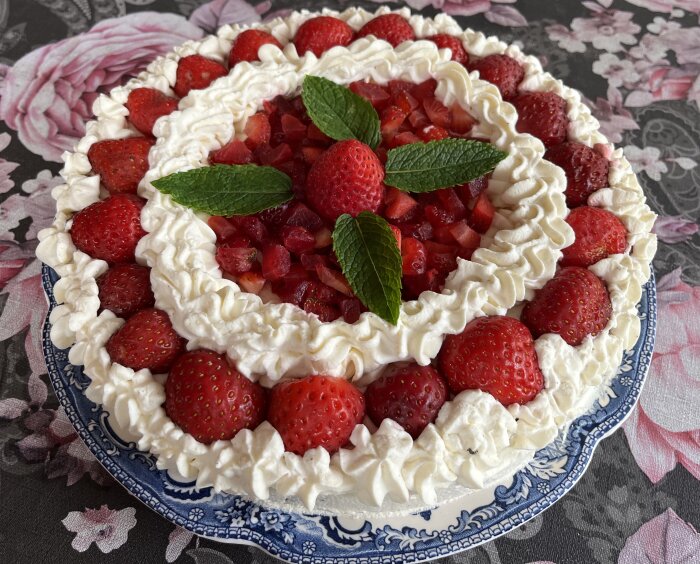 Gräddtårta med jordgubbar och fyra myntablad ovanpå, serverad på ett blåvitt porslinsfat med blommig duk.