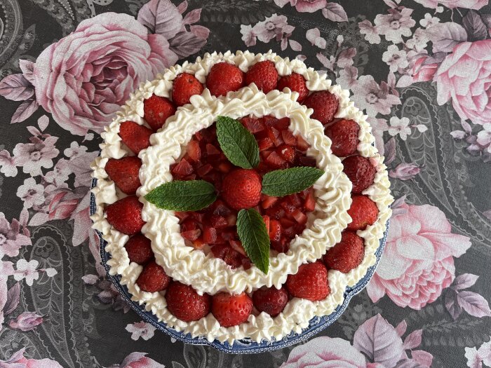 Jordgubbstårta med grädde och mintblad på ett blommigt bord.