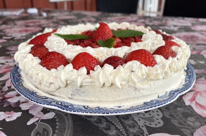 Gräddtårta med jordgubbar och myntablad på ett mönstrat fat, serverad på ett blommigt bord.