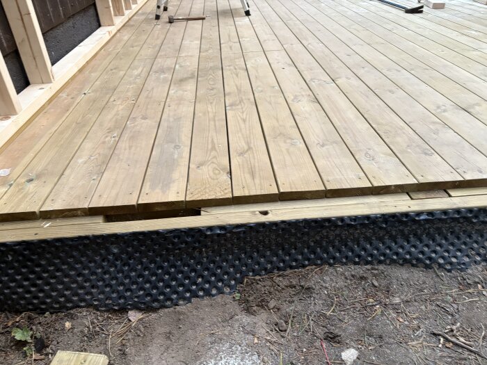 Trädäck vid kallförråd under uppbyggnad med tydlig golvås och fundament av syll i trä, skyddad av svart dräneringsplast mot marken.