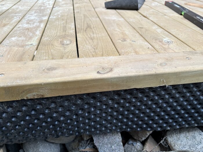 Bygger golv för kallförråd med träplankor, synlig syll och tätskikt. Stenfyllning under konstruktionen. Träverktyg i bakgrunden.