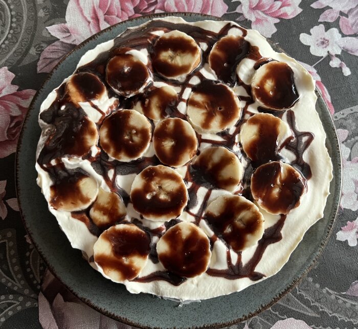 Bananoffeepaj med skivor av banan och chokladtopping på tallrik med rosa blommönster i bakgrunden.