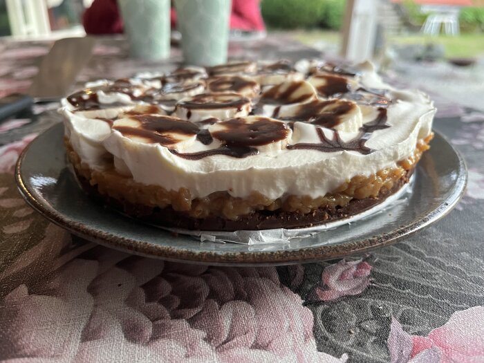 Bananoffe paj med botten av Domino salty caramel, toppad med skivad banan, vispgrädde och chokladsås på ett blommigt bord.