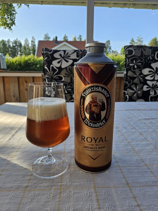 En stor ölburk av Franziskaner Weissbier Royal och ett glas öl stående på en bordsduk utomhus.