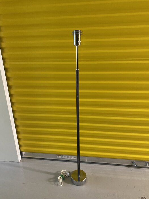 Gammal golvlampa utan lampskärm, med metallstomme och kabeln på basen, står framför en gul randig bakgrund.