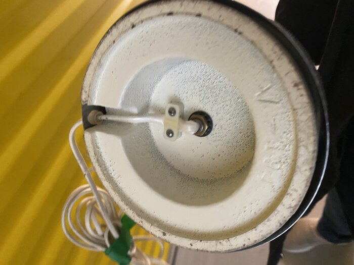 Närbild av undersidan på en gammal golvlampa med synlig kabel och strömbrytare, mot en gul bakgrund.